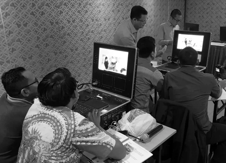 A group of men sit and stand around computer monitors displaying images. The atmosphere is focused and professional in a patterned-wall room.