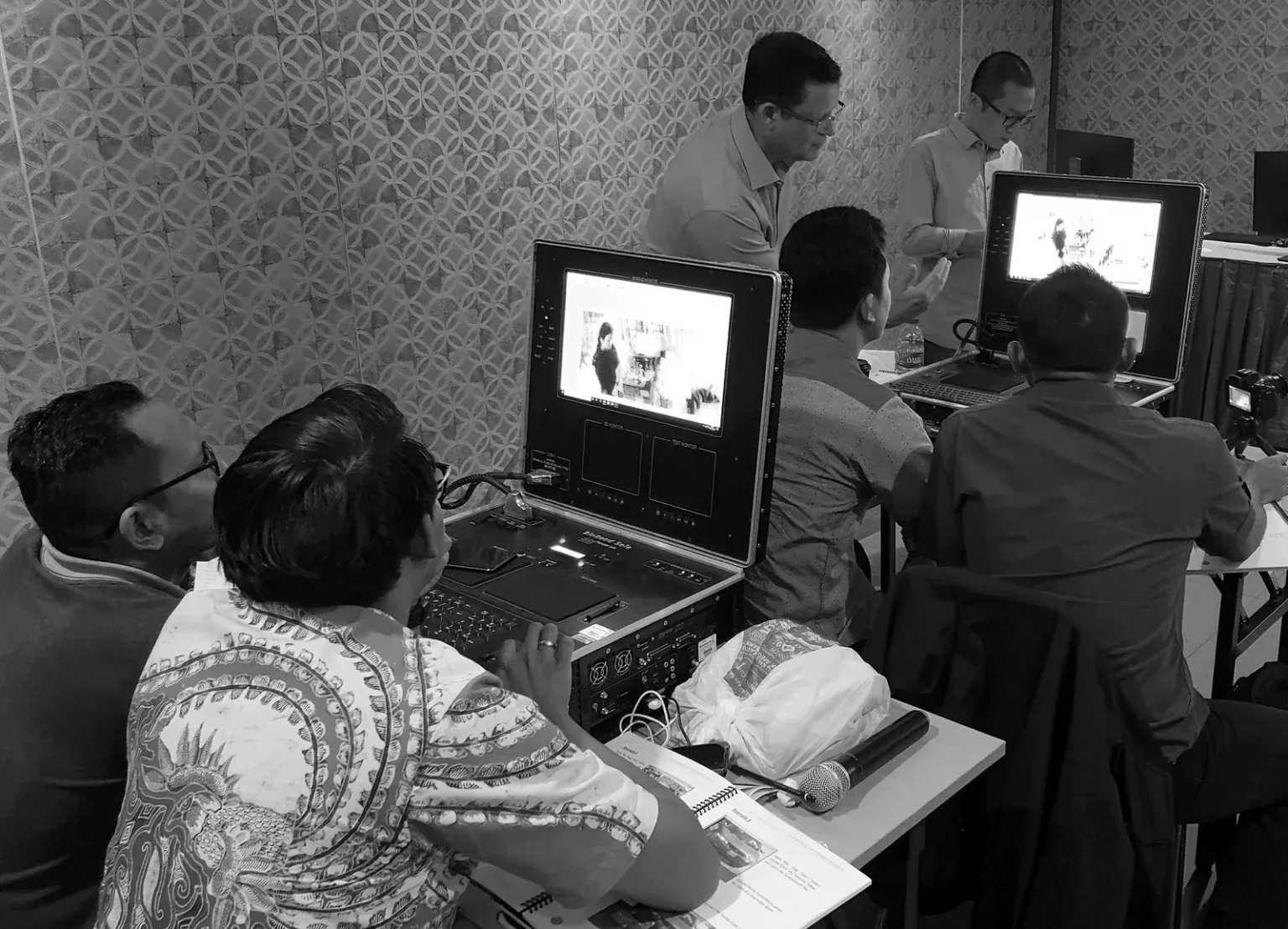 A group of men sit and stand around computer monitors displaying images. The atmosphere is focused and professional in a patterned-wall room.
